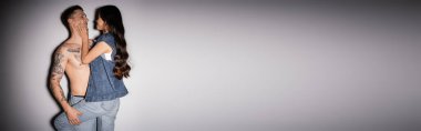 stylish asian woman in denim clothes touching face of shirtless man with tattooed body on grey background, banner