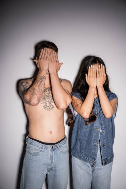 brunette woman in denim vest and shirtless man with tattooed body obscuring faces with hands on grey background