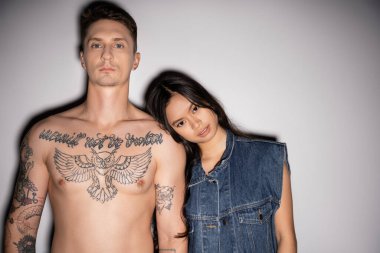 front view of shirtless tattooed man and asian woman in denim vest looking at camera on grey background