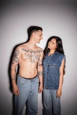 shirtless tattooed man and asian woman in denim clothes smiling at each other on grey background