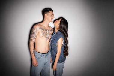 long haired asian woman in denim outfit sticking out tongue near smiling tattooed man on grey background