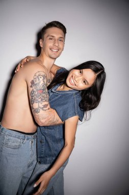 cheerful tattooed man and young asian woman in denim outfit smiling at camera on grey background