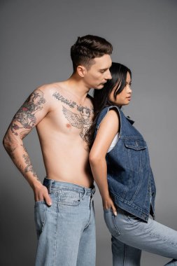 sensual asian woman and shirtless man with tattooed body posing with hands in back pockets of jeans on grey background