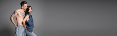 shirtless tattooed man and trendy asian woman posing with hands in back pockets of jeans on grey background, banner