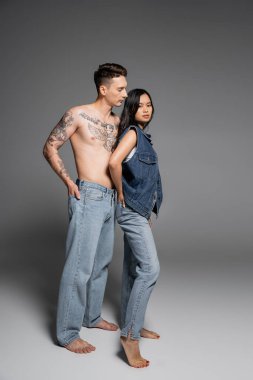full length of barefoot tattooed man in jeans near brunette asian woman in denim outfit posing on grey background