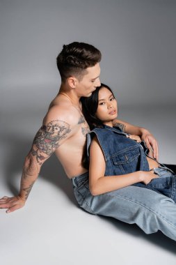 pretty asian woman in denim vest looking at camera while sitting near shirtless tattooed man on grey background