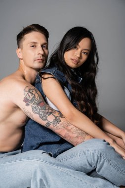 shirtless tattooed man in jeans and asian woman in denim outfit looking at camera while sitting on grey background