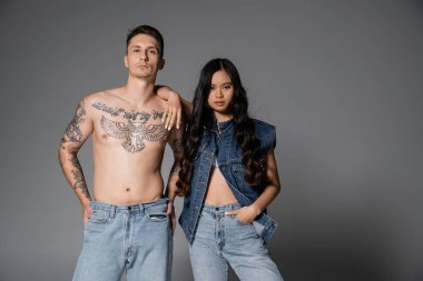 stylish asian woman in denim clothes and shirtless man with tattooed body looking at camera on grey background