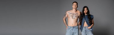 shirtless man with tattooed body and brunette asian woman in denim clothes posing on grey background, banner