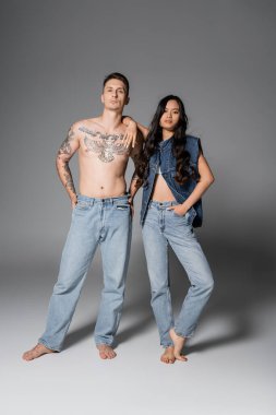 full length of trendy asian woman and shirtless man with tattooed body posing in jeans on grey background clipart