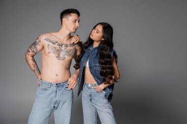 asian woman in stylish denim outfit looking at shirtless tattooed man in jeans on grey background