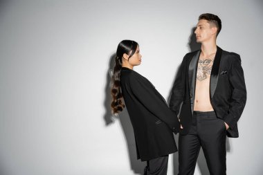 long haired asian woman in stylish attire looking at tattooed man in black blazer on grey background