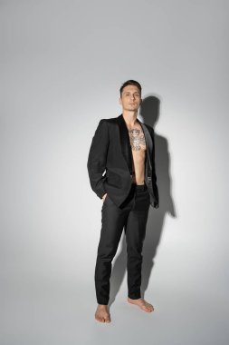 full length of barefoot man with tattooed body standing with hand in pocket on grey background with shadow