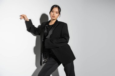 sensual asian woman in black elegant outfit looking at camera on grey background with shadow