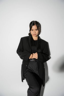 brunette asian woman in black elegant suit looking at camera on grey background