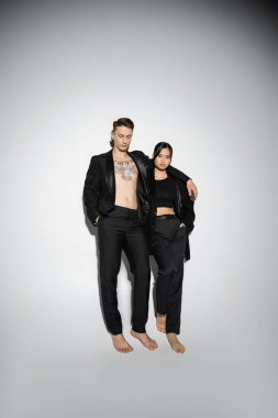 full length of barefoot interracial couple posing in black elegant pantsuits on grey background