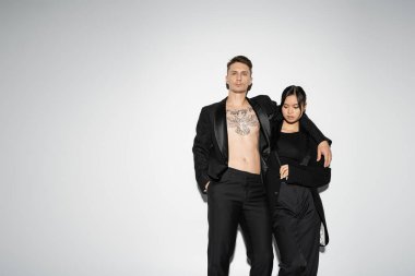 tattooed man in black trendy suit embracing elegant asian woman while posing with hand in pocket on grey background