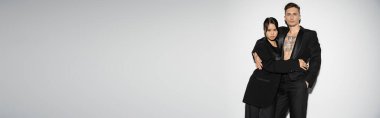 brunette asian woman and tattooed man wearing black stylish suits and embracing on grey background, banner