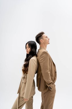 side view of interracial couple in beige suits standing back to back isolated on grey