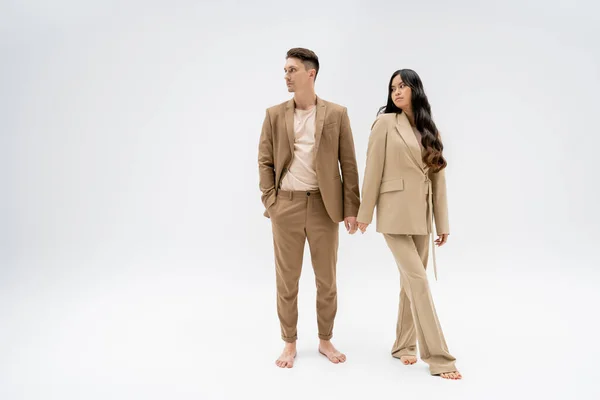 full length of barefoot interracial couple in beige pantsuits looking away on grey background