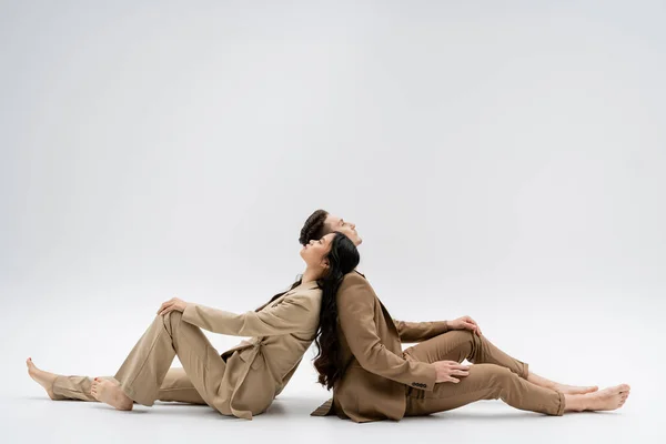 stock image side view of multiethnic couple in beige suits sitting back to back on grey background