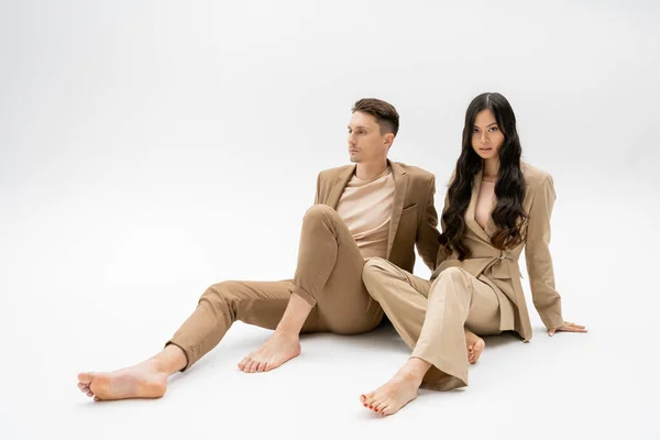 full length of barefoot interracial couple in beige suits sitting on grey background