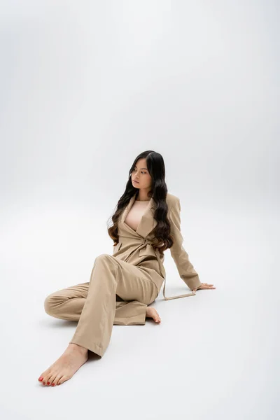 full length of barefoot asian woman with long brunette hair sitting in beige pantsuit on grey background