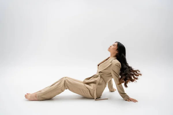 side view of barefoot asian model with long hair sitting in fashionable pantsuit on grey background
