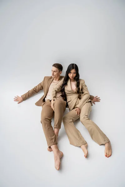 full length of barefoot multiethnic couple in fashionable outfit sitting on grey background