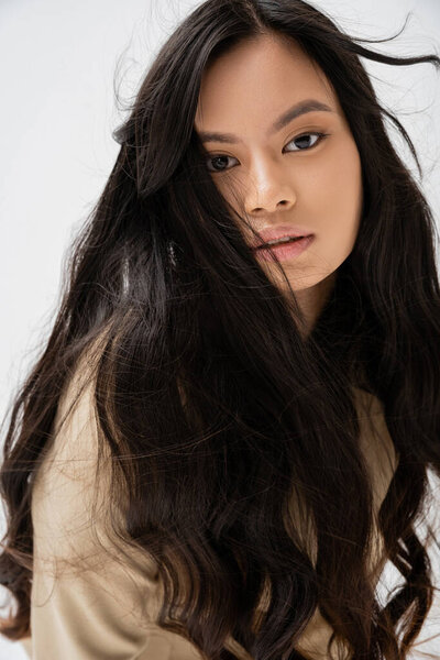 portrait of brunette asian woman with long hair and natural makeup looking at camera isolated on grey