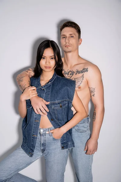 shirtless tattooed man and asian woman in denim outfit looking at camera while posing on grey background