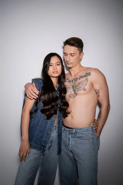 long haired asian model in denim clothes looking at camera near shirtless man with tattooed body on grey background