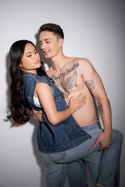 smiling tattooed man embracing sensual asian woman in denim outfit on grey background