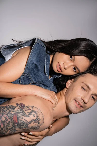muscular and tattooed man piggybacking sensual asian woman looking at camera on grey background