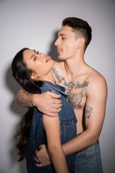 brunette asian woman in denim vest and shirtless man with tattooed body sticking out tongues on grey background
