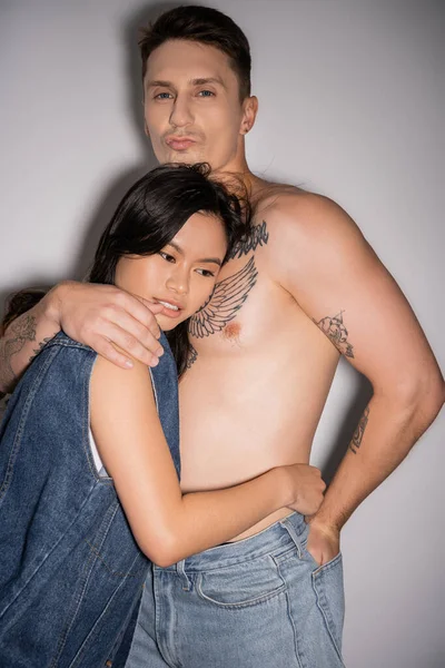 tattooed and muscular man pouting lips and hugging asian woman in denim vest on grey background