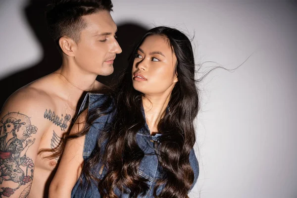 shirtless tattooed man and long haired asian woman in denim vest looking at each other on grey background