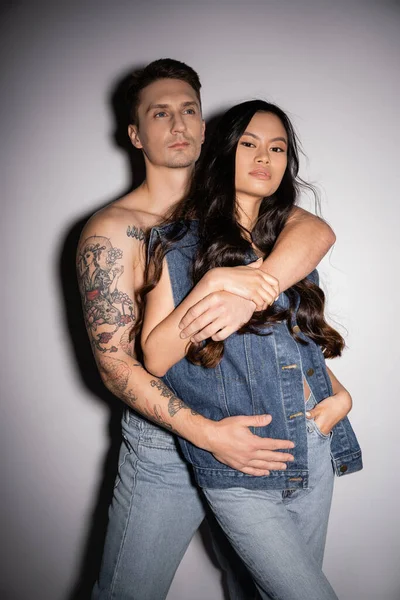 shirtless tattooed man hugging brunette asian woman in denim clothes looking at camera on grey background