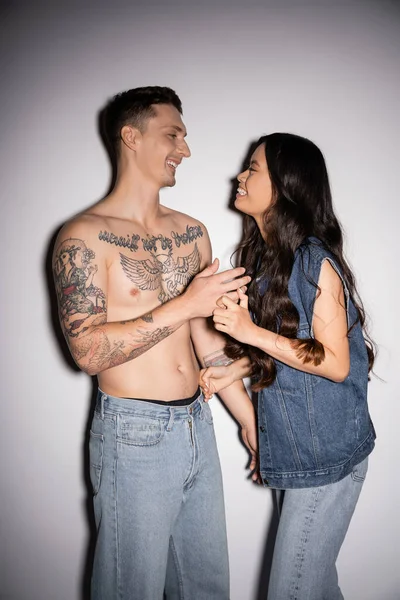 cheerful asian woman and shirtless tattooed man smiling at each other on grey background