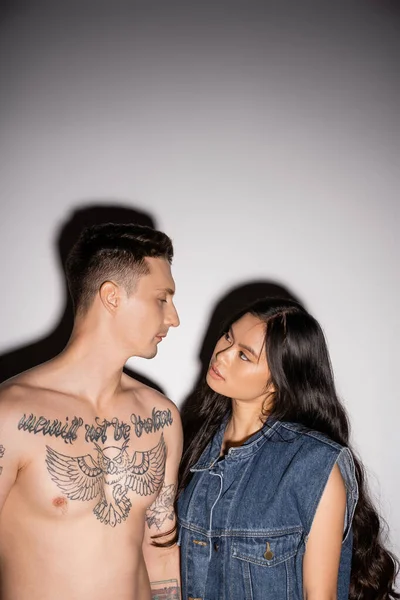 shirtless man with tattooed body and brunette asian woman with long hair looking at each other on grey background
