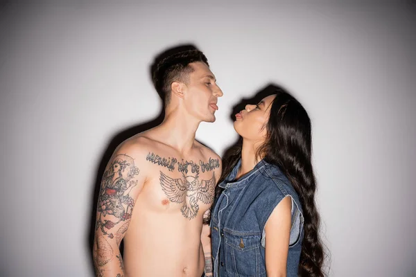 stock image shirtless tattooed man and asian woman in denim clothes sticking out tongues on grey background