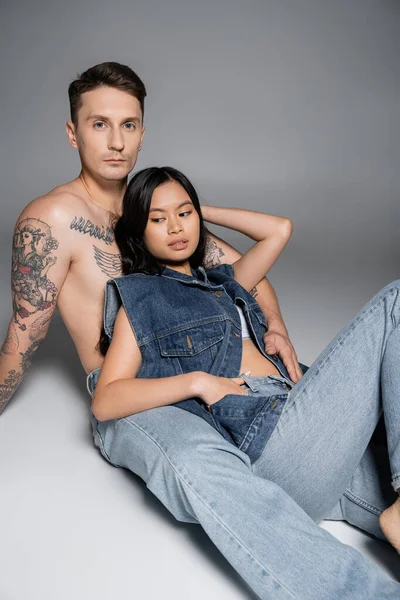 tattooed shirtless man looking at camera while sitting near asian woman in denim clothes on grey background