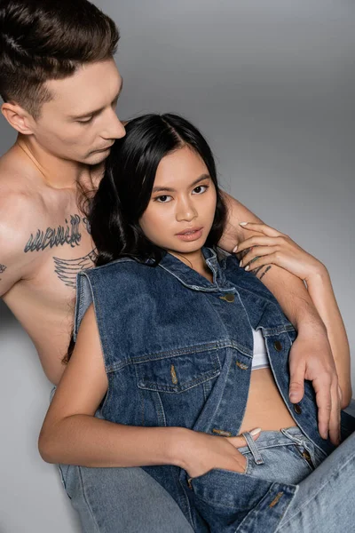 high angle view of young asian woman in denim vest looking at camera near shirtless tattooed man on grey background
