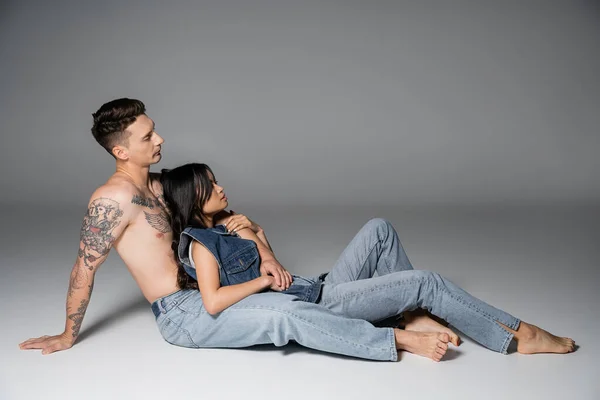 side view of brunette asian woman and shirtless tattooed man in jeans sitting on grey background