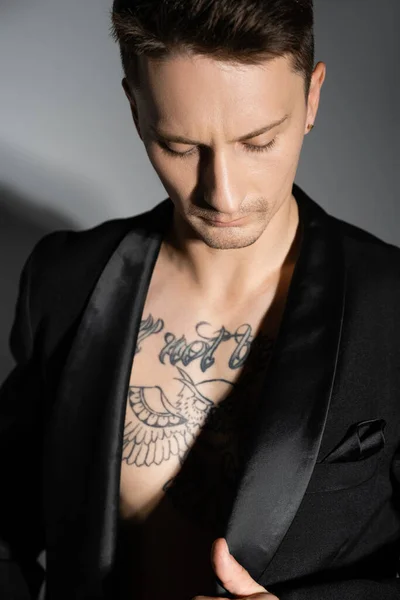 trendy man with tattooed chest wearing black blazer on grey background