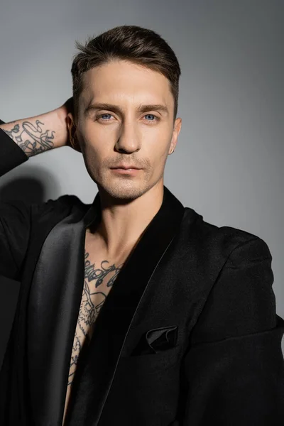 stylish tattooed man in black blazer posing with hand behind head while looking at camera on grey background