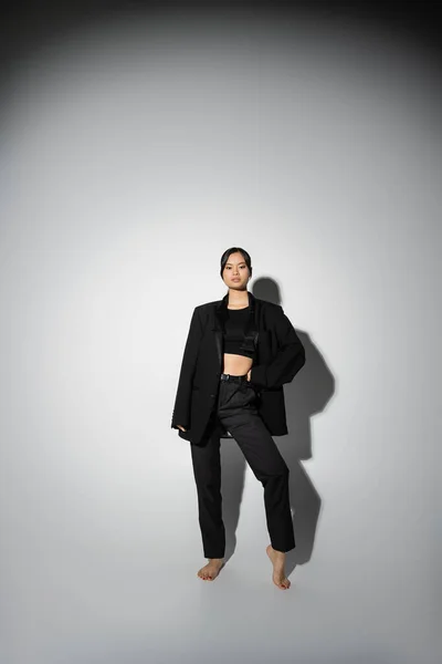 stock image full length of barefoot asian woman in black elegant attire posing with hand on waist on grey background with shadow