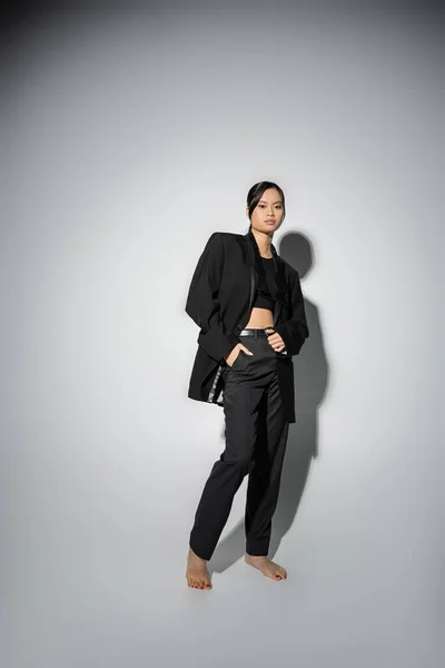 full length of barefoot asian woman in black pantsuit posing with hand in pocket on grey background with shadow