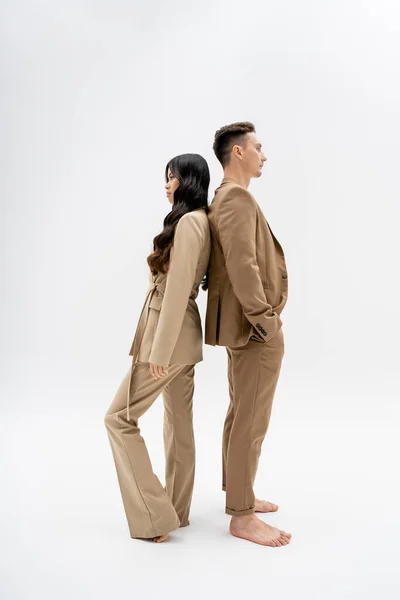 side view of barefoot multiethnic couple in stylish pantsuits standing back to back on grey background