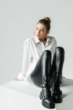 full length of young woman in stylish outfit sitting on white cube and looking away isolated on grey clipart